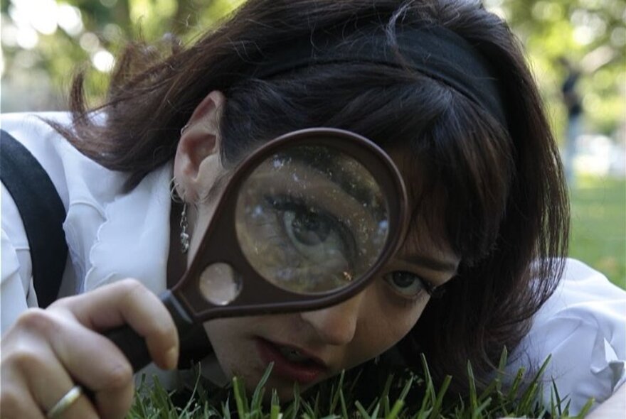 A woman laying on grass with one eye peering through a magnifying glass