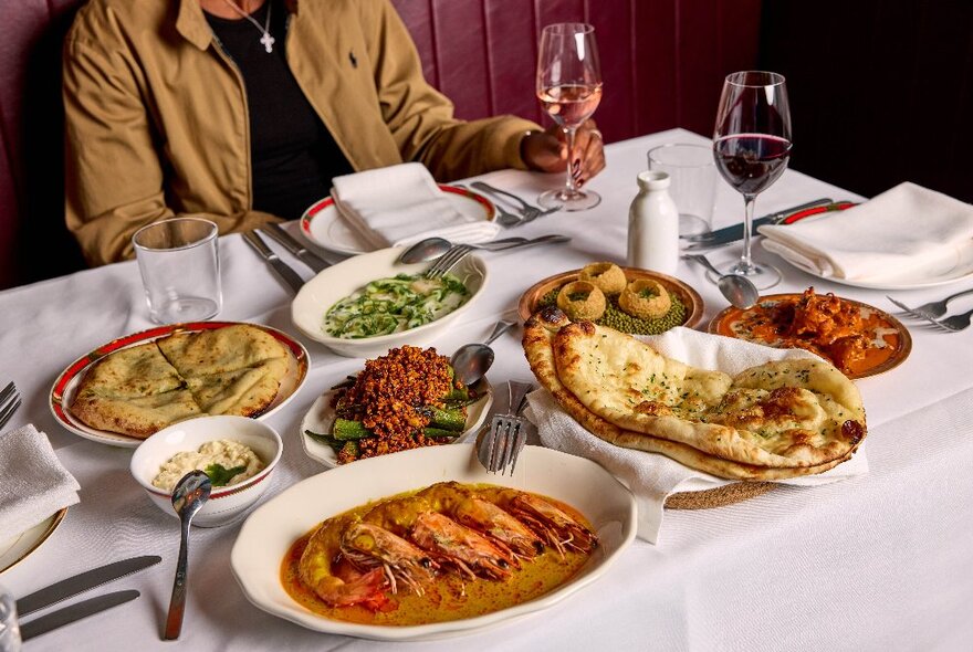 A restaurant table with plates of Indian food arranged on it, two glasses of wine, cutlery, cloth napkins, and a person seated on a banquette behind the table.
