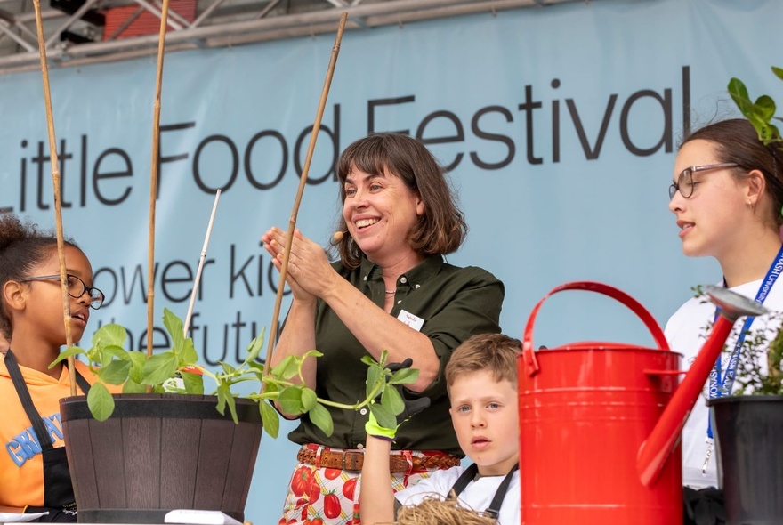 An adult and a few primary-school aged children standing at a table that holds a potted plant and a red watering can, with slightly obscured signage in the background that reads 'Little Food Festival'. 