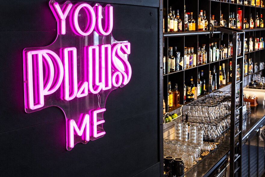 A bar with shelves of spirits on display and a pink neon sign that says, You Plus Me.
