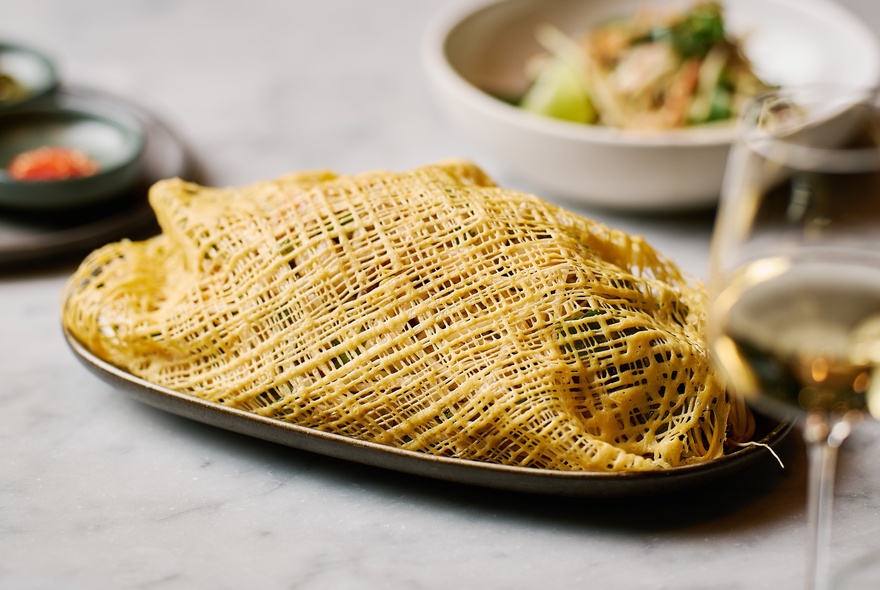 Thai dish in a mesh omelette nest on a table with other dishes and glass of white wine.