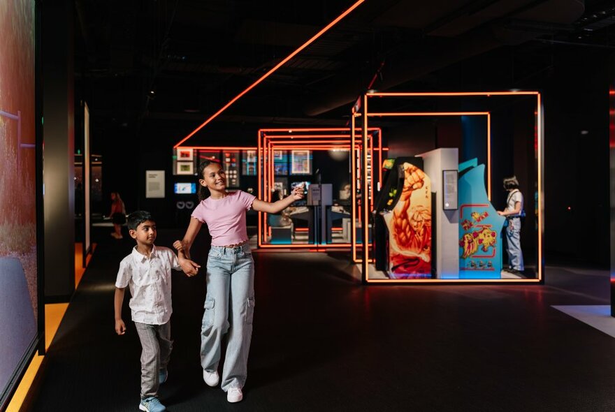 Two kids wander through an exhibition space that's dimly lit with cubes of neon light around displays. 