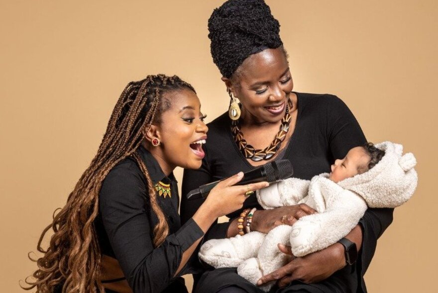 Two women with a baby against a tan background, the baby wears a hooded furry onesie.