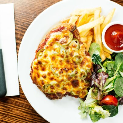 Overhead view of a chicken parma, fries and salad on a white plate, with cutlery on a napkin alongside.