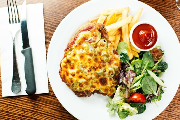Overhead view of a chicken parma, fries and salad on a white plate, with cutlery on a napkin alongside.
