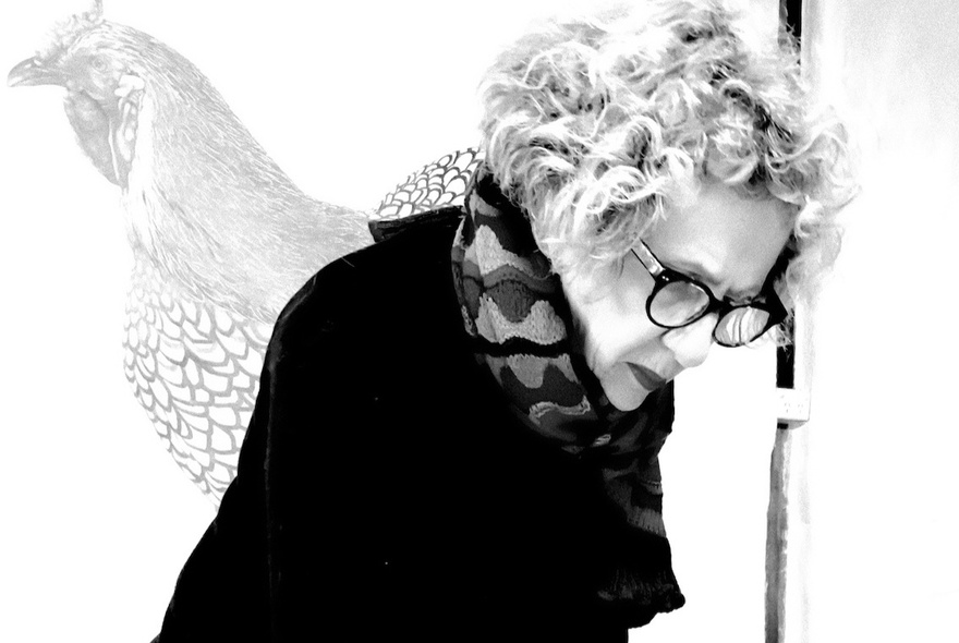 Black and white photograph of a woman with voluminous, curly hair and thick-rimmed glasses, leaning forward with her head tilted down.