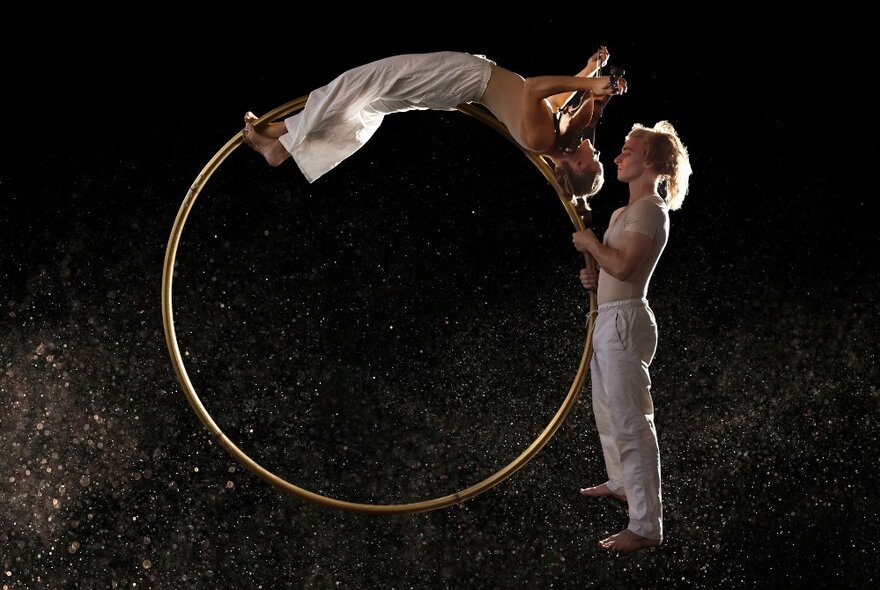 Two performers on a darkened stage, one holding a huge metal ring while the other lays across the top of it, their body curved into the structure.