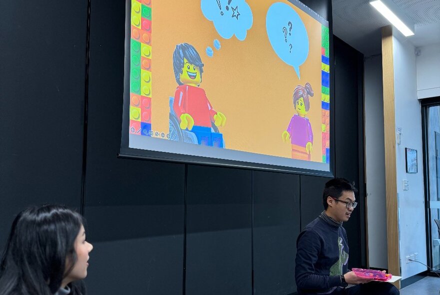 Two people seated under a large screen in a workshop venue.