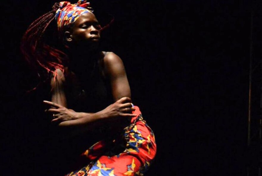African dancer in darkly lit room, wearing colourful red sarong and headcovering.
