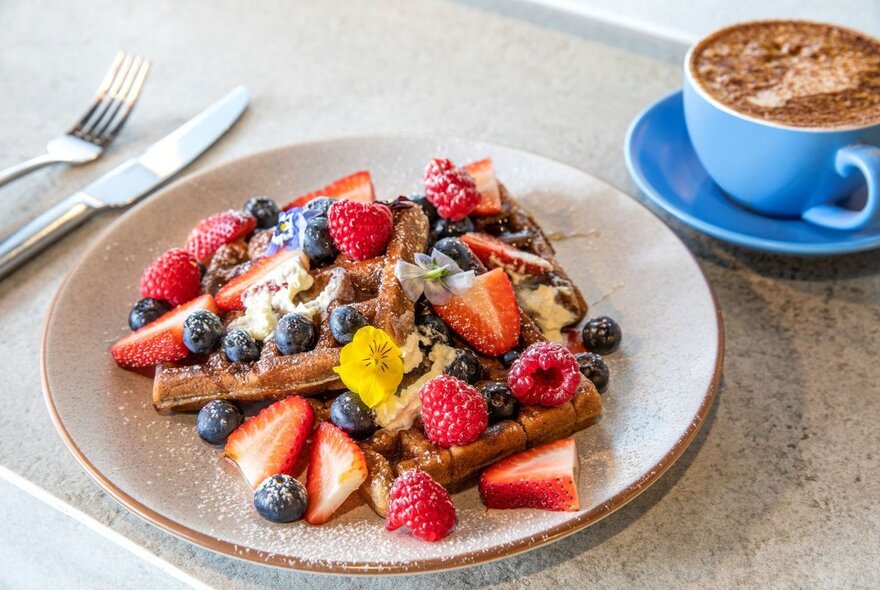A plate of breakfast waffles with fresh fruit, with cutlery and a cup of coffee.