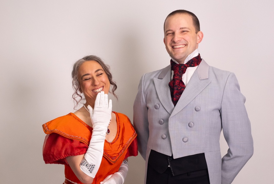 Two impro actors in Regency period costumes, the woman bringing a gloved hand up to partly cover her grin.