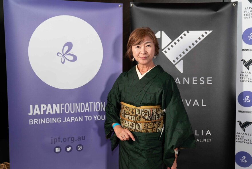 A Japansese woman in a kimono in front of two promotional banners.