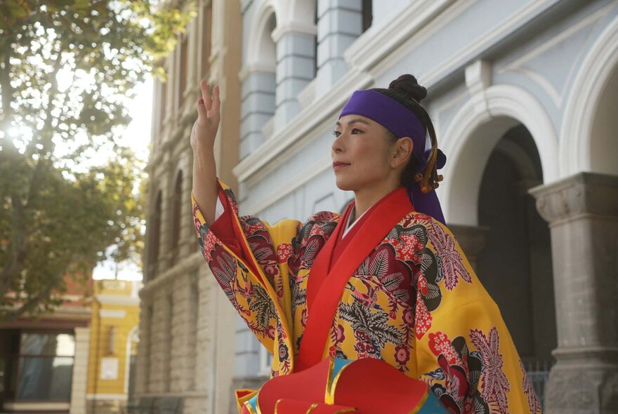 Woman wearing a bright kimono with her arm raised, standing outside an arched portico.