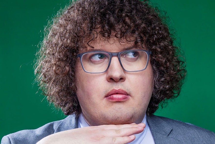 Comedian Ray O'Leary looking upwards to his left with a hand flat under his chin, against a green background. 