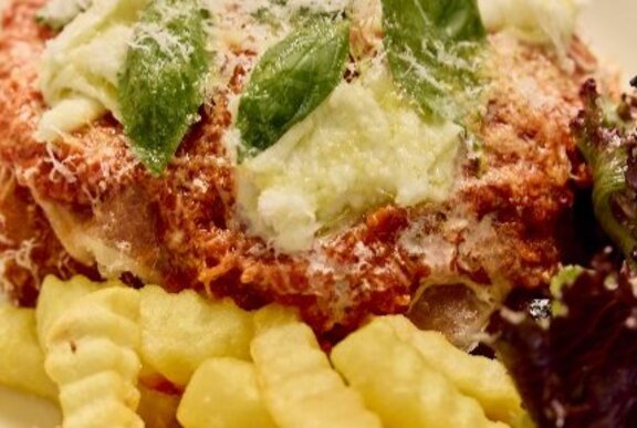 Close up of a chicken parmagiana with tomato sauce, cheese and basil leaves on top, next to crinkle-cut fries.