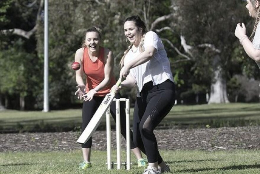 Women playing cricket, one woman about to hit a ball, another leaning forward to catch the ball.