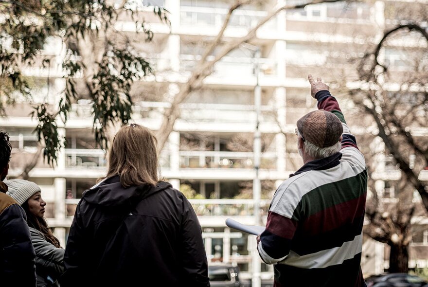 A tour guide leading a walking street tour, pointing to a large apartment block in the background, with a few people standing around listening.