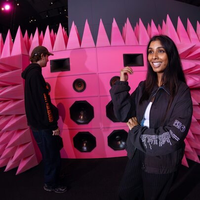 Two people standing in front of a large pink spiky wall of speakers in an exhibition space.