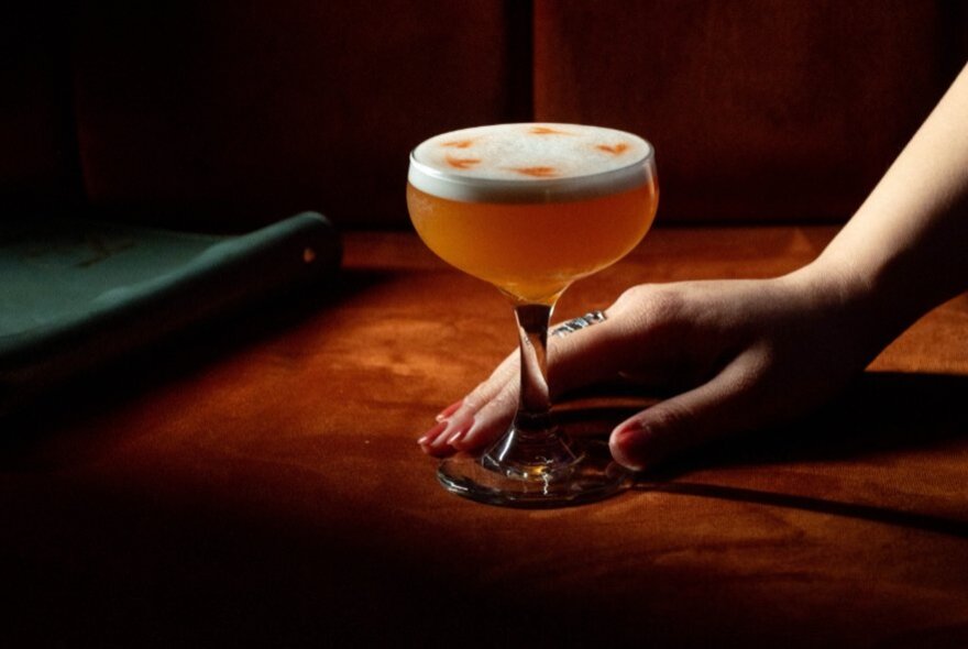 Hand holding an orange cocktail with frothy top.