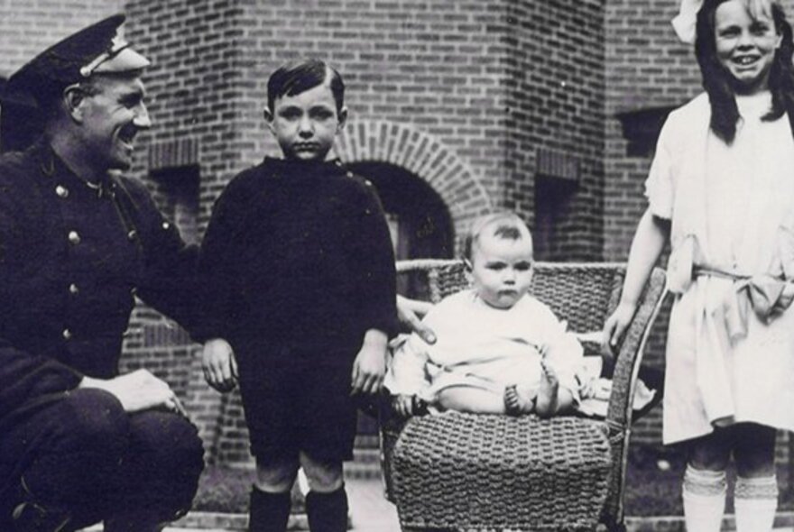 Two young children, a baby in a chair and a man in uniform, in a black and white vintage family photo wearing clothes dating from the 1920s.