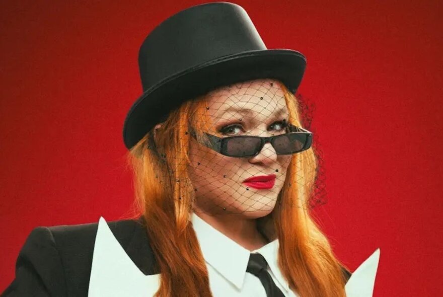 A woman wearing a black top hat with black veil over her face, dark sunglasses and a black suit and tie.