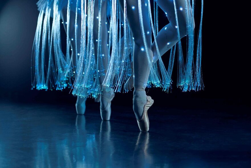 Ultraviolet-lit legs of ballet dancers pirouetting in a row with dangling strands of light.