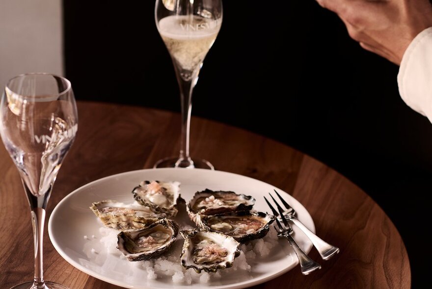 A round wooden table with a plate of oysters on rock salt and two dessert wine glasses, one being poured. 