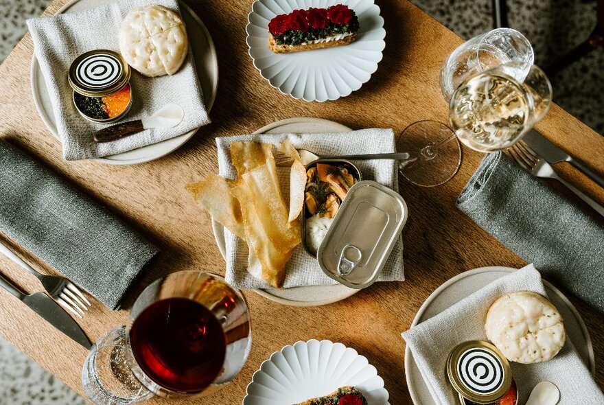 A wooden table laden with gourmet delicacies, wide glasses, napkins and cutlery, seen from above.