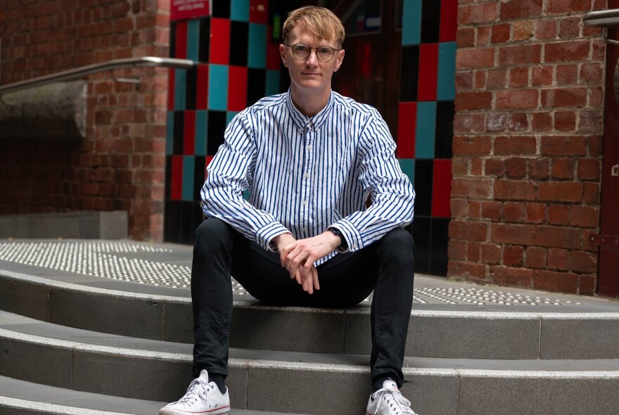 Artist and academic Drew Pettifer, sitting on the steps outside a tiled and red brick entrance to a building.