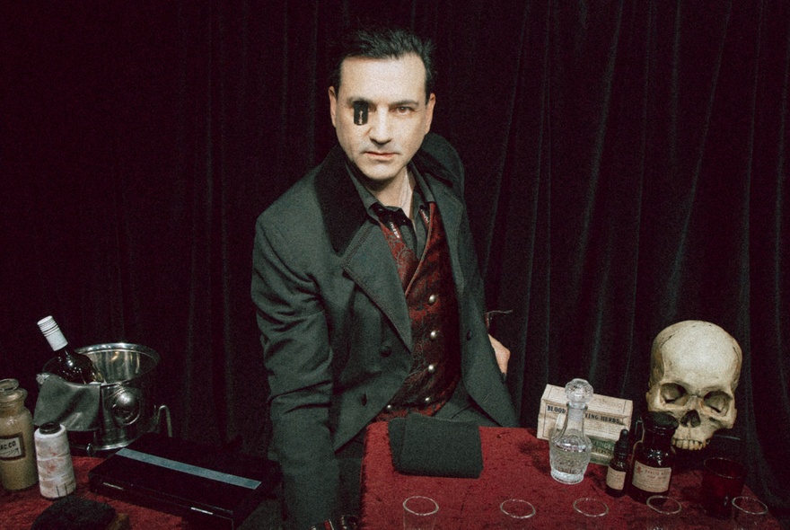 Magician with one eye hidden, surrounded by alchemical paraphernalia incuding skull, glass vials and bottles.