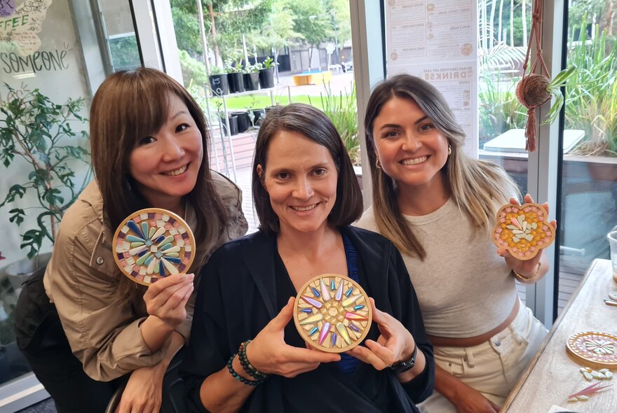 Three workshop participants smiling and holding up multicoloured mosaic tiled coasters, seated at a workbench.