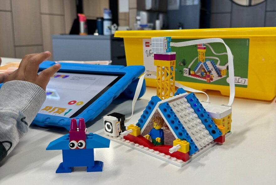 A child's hand touching a tablet with a Lego toy model of a house and other Lego toys.