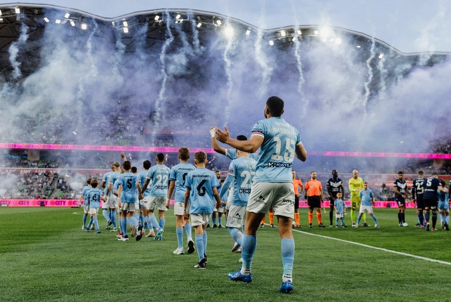 A mens soccer team in pale blue kit entering a soccer field while fireworks go off.