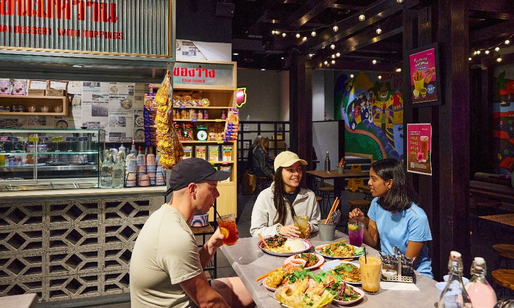 Three young people enjoying various dishes in a colourful, hawker-style Thai restaurant.