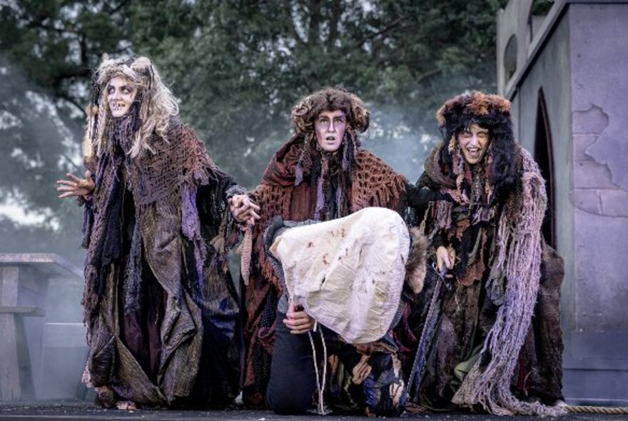 Actors dressed as the ragged witches from the play Macbeth, performing outdoors in the gardens at twilight.