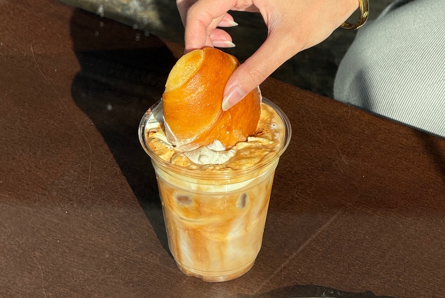 Hand dipping a Japanese milk bun into a frothy coffee drink.