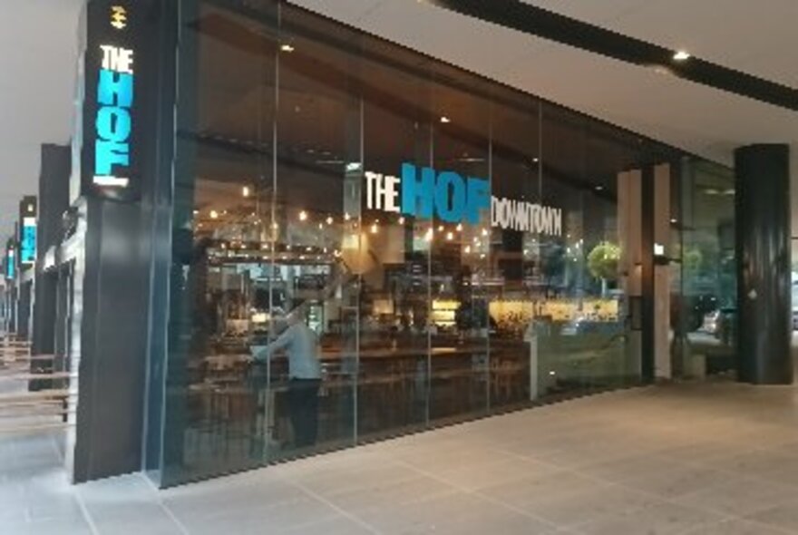 The glass wall exterior of The Hof Downtown bar and cafe, with signage and patrons inside.