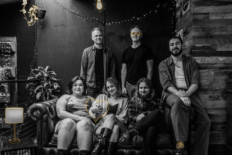 Group of musicians, some seated on a chesterfield, the others standing behind them, in a space decorated with plants and swags of lights.