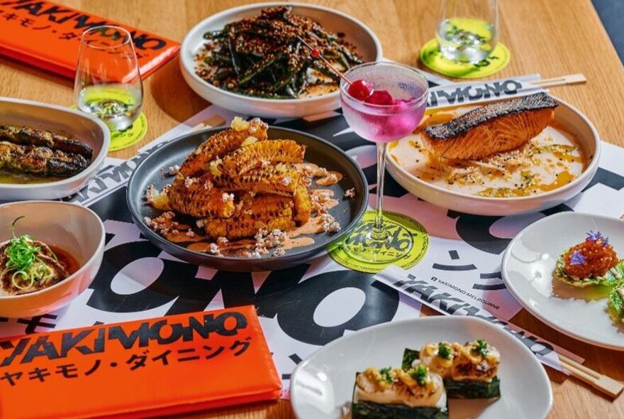 Several plates of Japanese food arranged on a restaurant dining table, with a pink cocktail and menus.