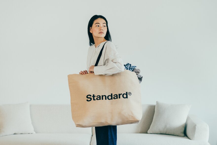 A person carrying a large tote shopping bag with the word STANDARD printed on it, standing in front of a white sofa.