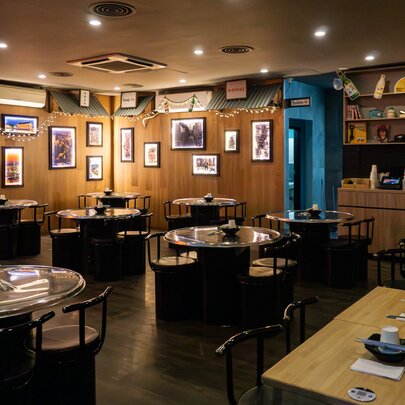 The interior of a restaurant with tables and chairs arranged in the space, small pictures hung on the wood panelled walls, and downlights.