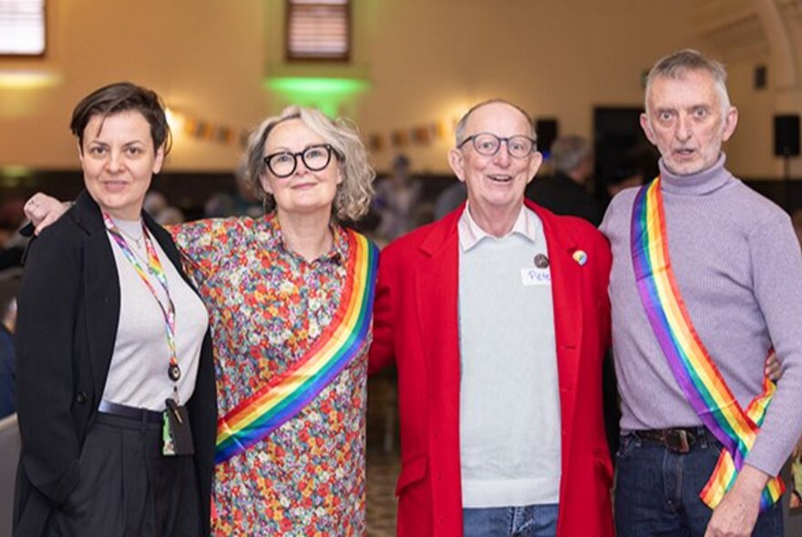 Older gay men and women standing with arms linked wearing rainbow sashes.