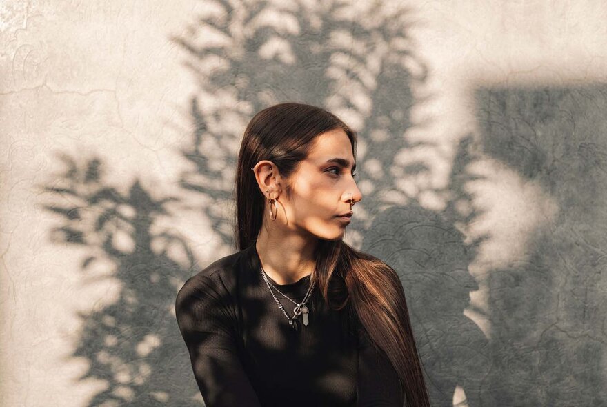 A woman in profile, her long hair pushed behind her ear, standing in front of a grey concrete wall with the shadow on a leafy tree casting patterns across her face and the wall.
