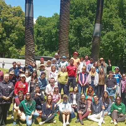 Large group of people sitting under the shade of a large tree in a park setting.