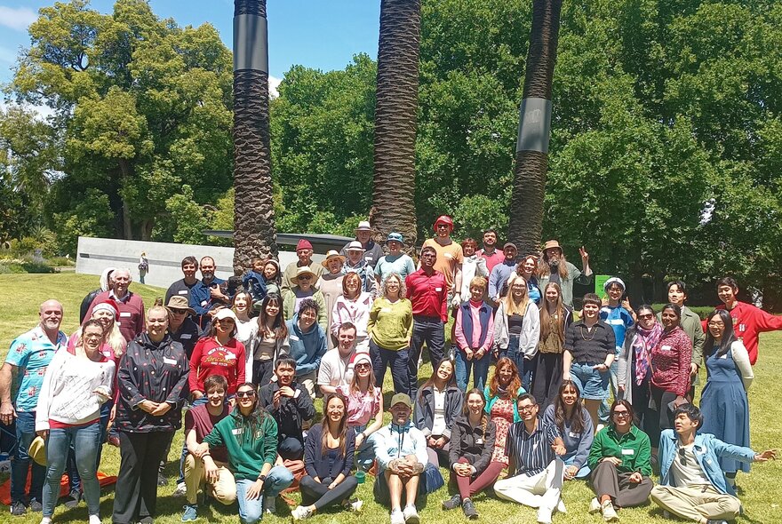 Large group of people sitting under the shade of a large tree in a park setting.