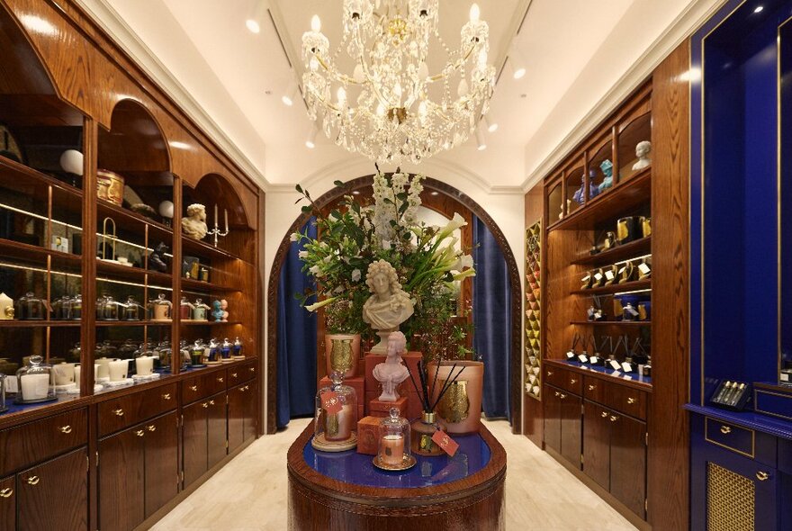 A glamorous candle shop with polished wooden shelving, an island display, a chandelier and blue velvet curtains.