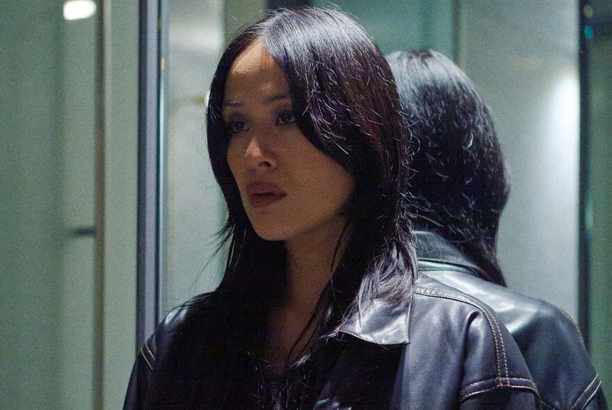 The head and shoulders of a woman with shoulder length black hair falling to her shoulders, her face angled to one side, wearing a black leather jacket, standing with a mirror behind her.