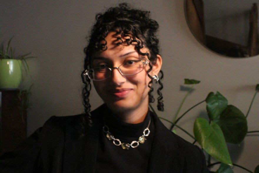 Young woman with curly hair and spectacles, wearing a silver necklace over a black jumper, plants in the background.