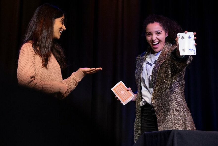 Two women on stage, on performing a magic trick involving playing cards facing the audience with a wide smile, the other looking on smiling in amazement. 