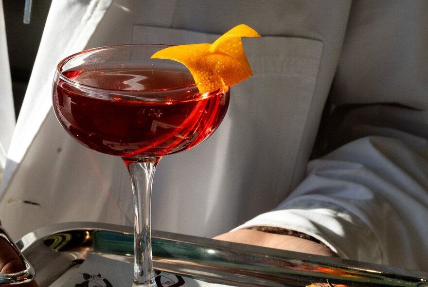 A red cocktail with an orange twist on a silver tray.